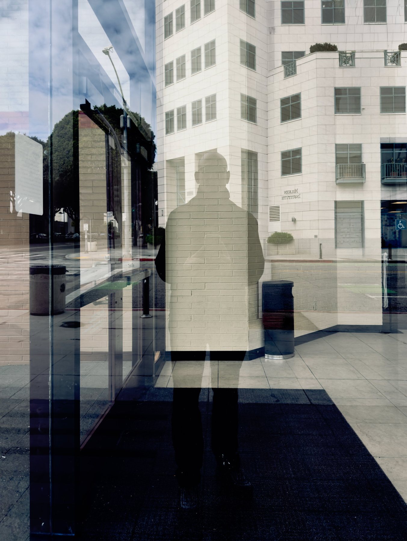 No End In Sight 139: The artist's pale reflection overlaying a white office building in a glass facade, with a rubbish bin and dark paving.