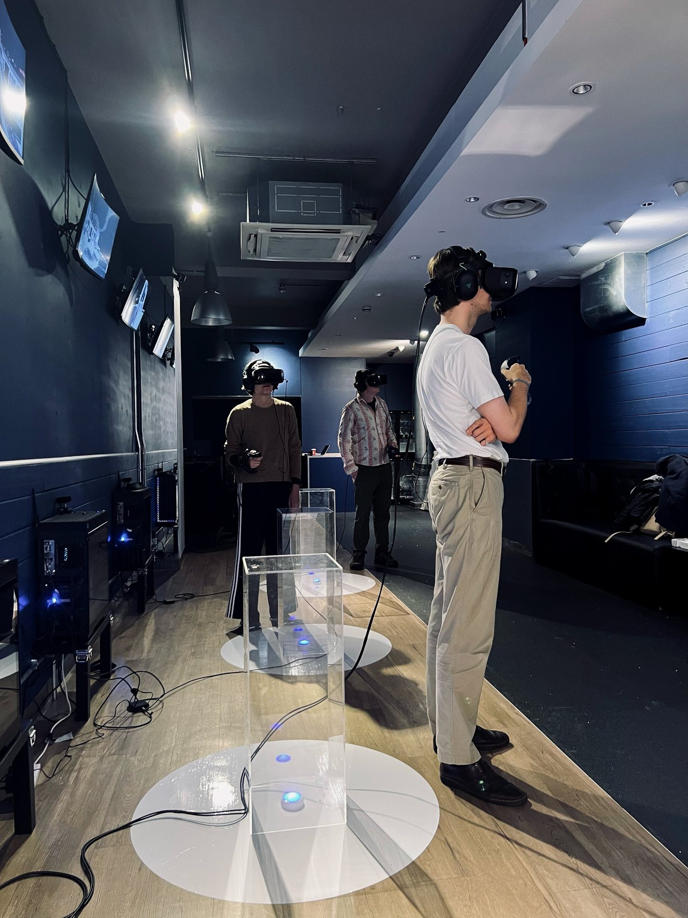 Visitors in headsets stand among clear plinths in the dark-blue gallery, one in a white t-shirt in the foreground.