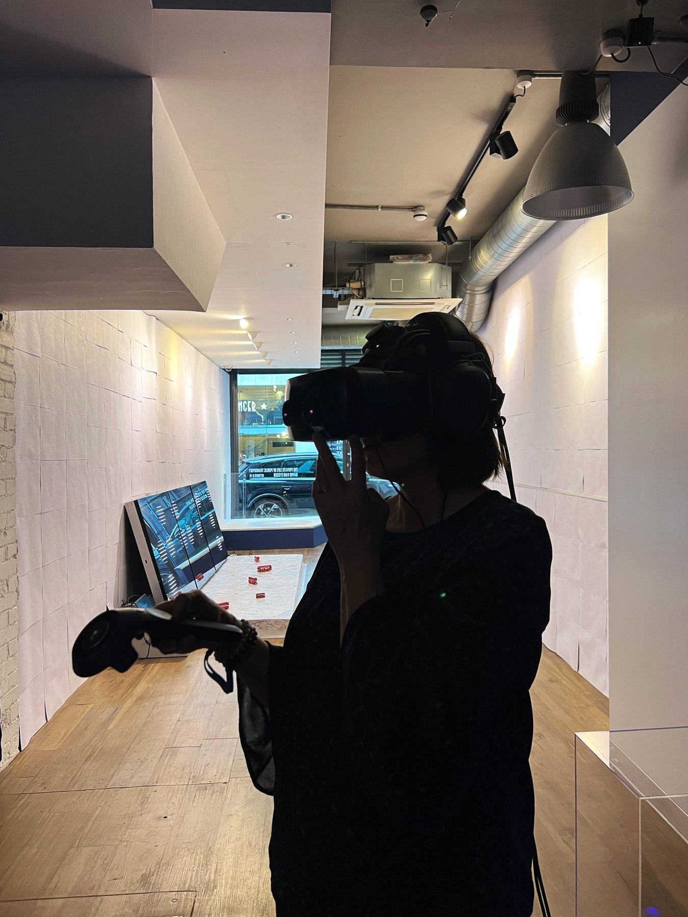 A woman in a headset holding a controller, silhouetted against the white gallery and a street window beyond.