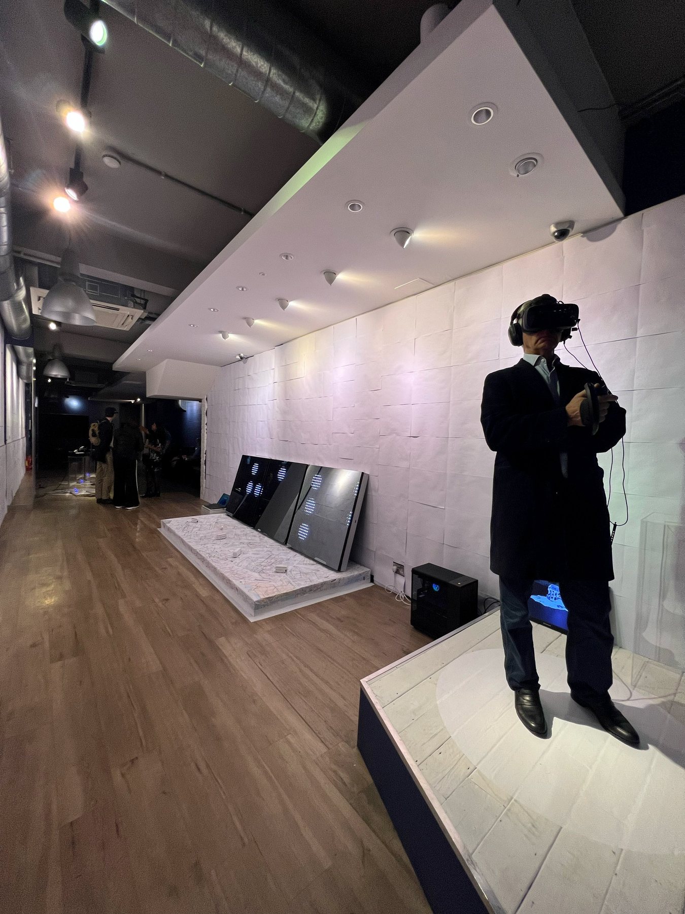 A man in a dark coat and headset on the white platform, with the braille-papered wall and map platform behind him.