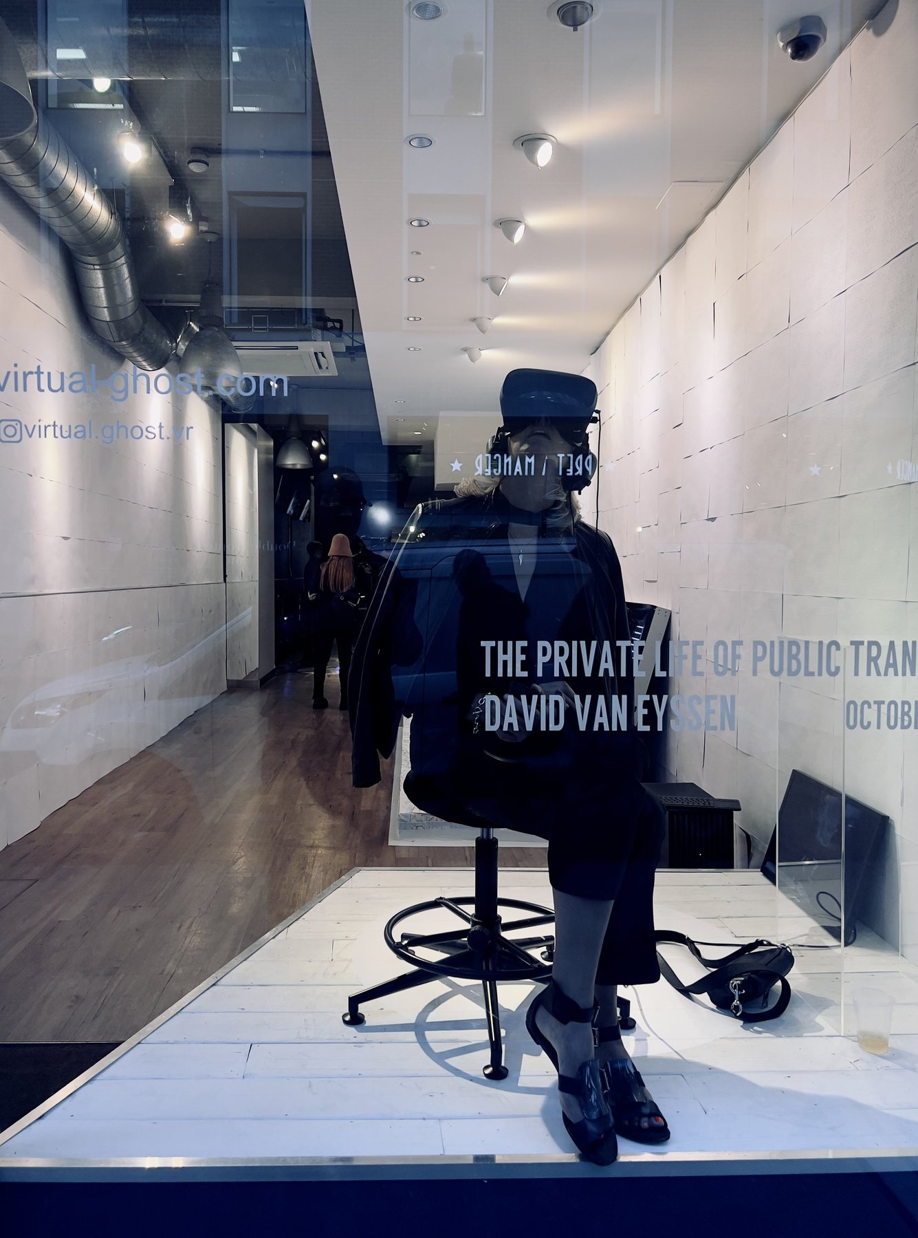A woman seated on a stool wearing a headset, reflected in the gallery window beside the exhibition title.