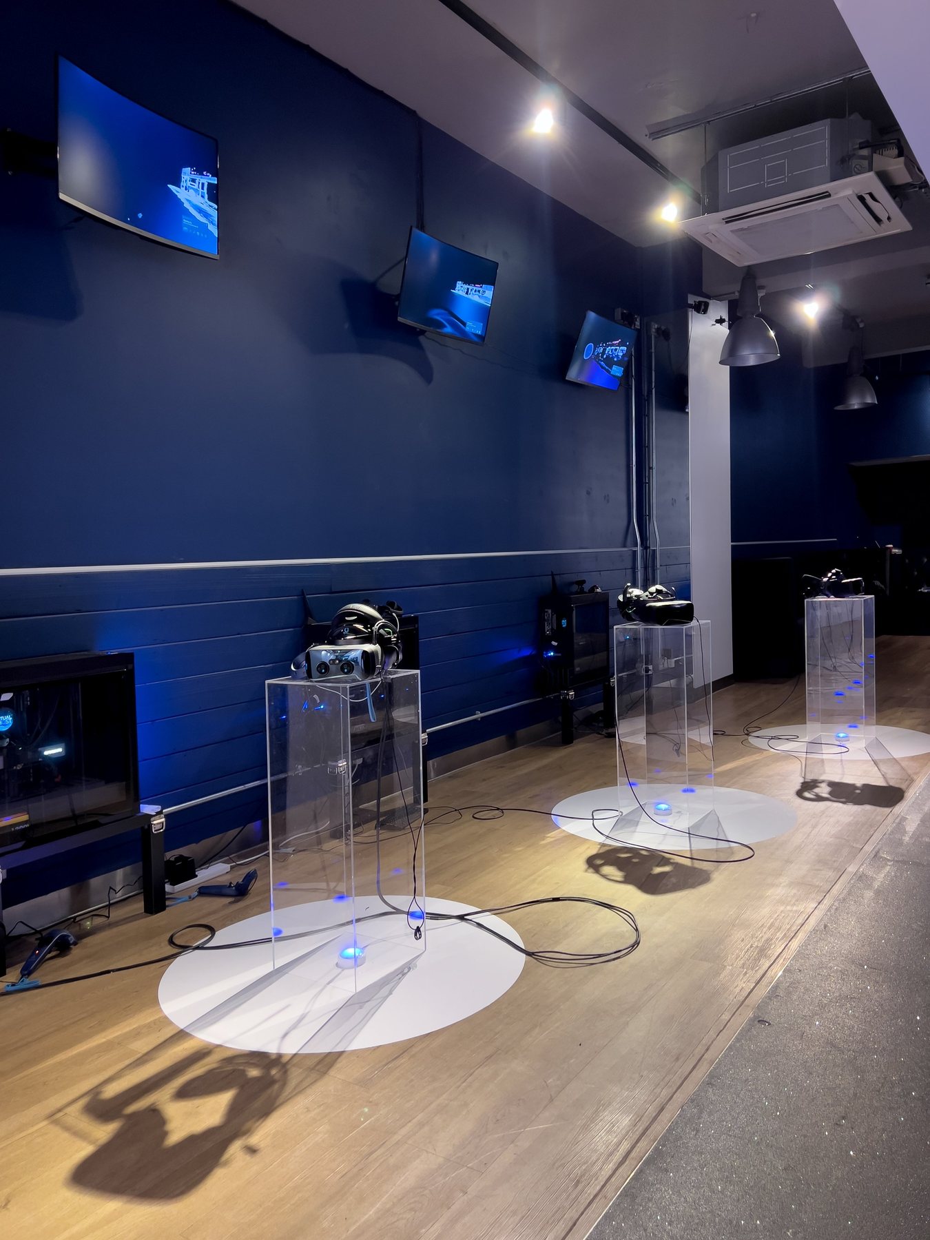 Three clear plinths with headsets on circular pools of light against a deep blue wall with screens.