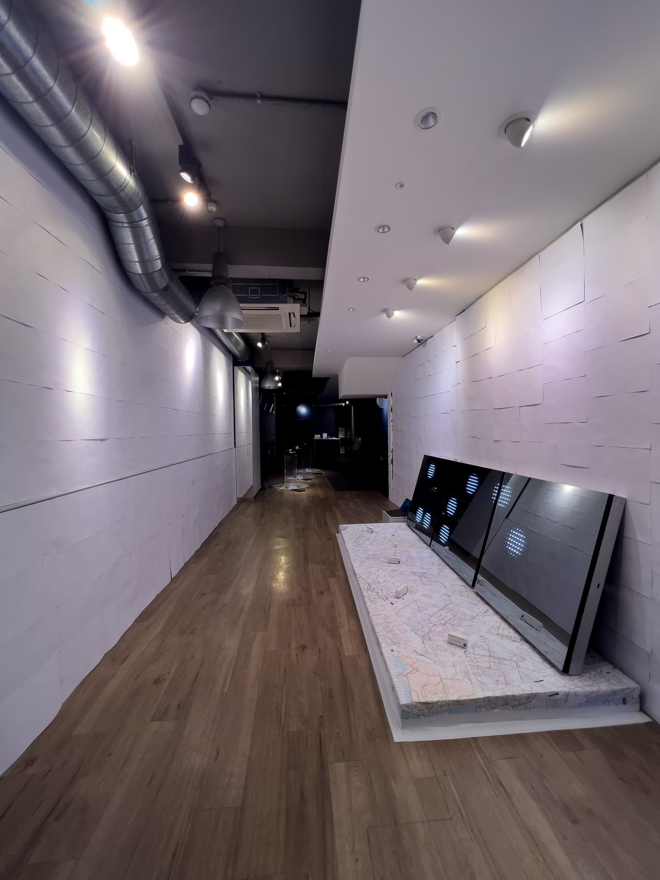 Narrow white gallery with braille wallpaper at left and angled mirror screens on a map-covered platform at the end.