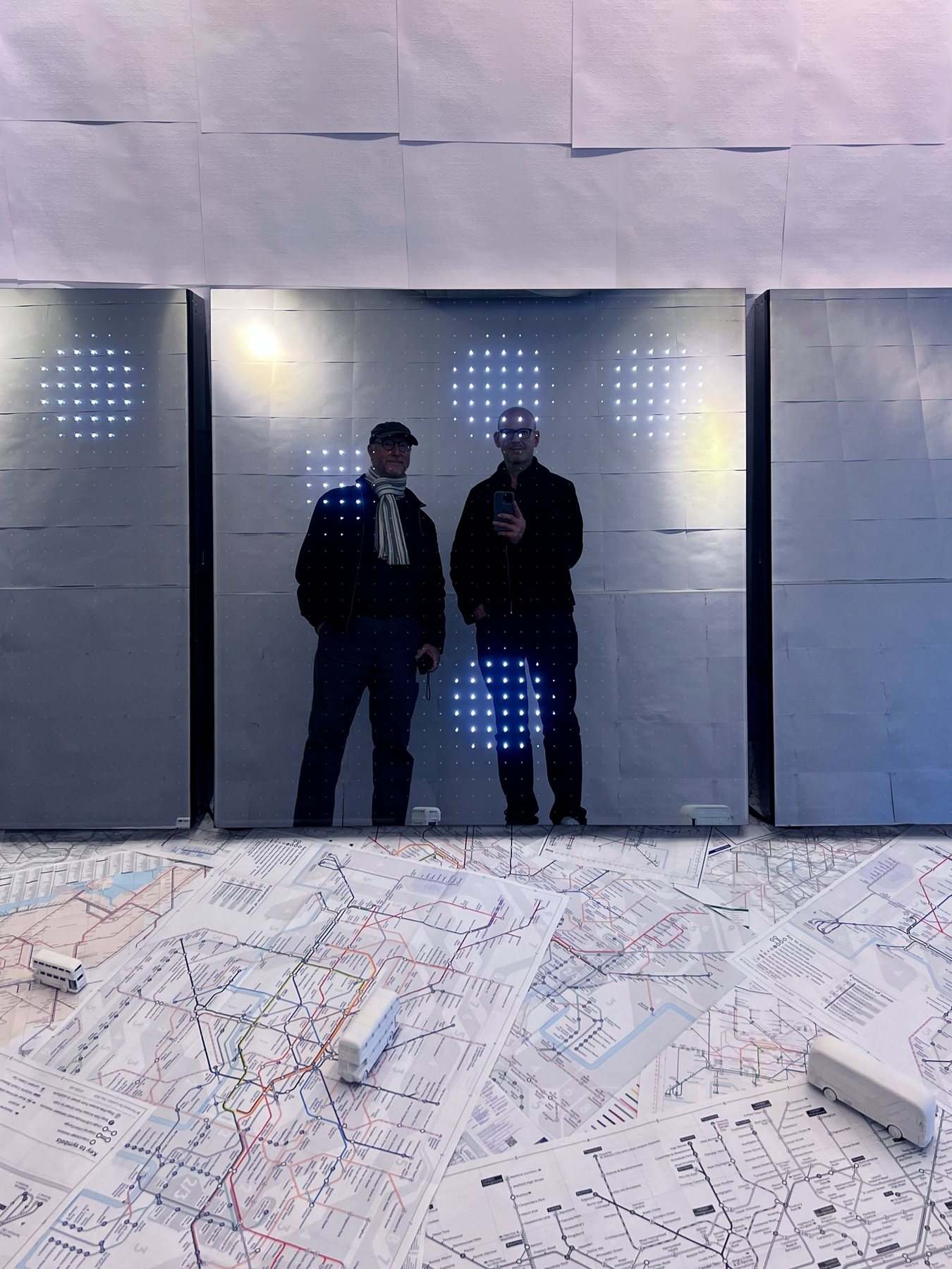 Two men reflected in a dark mirrored panel dotted with lights, above a floor map with miniature buses.