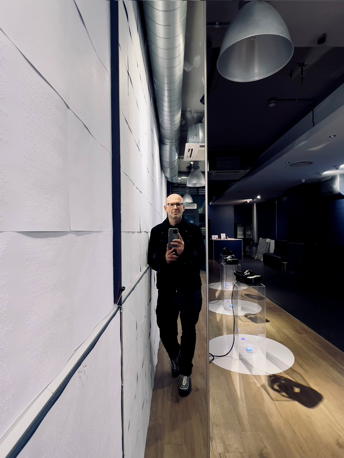The artist's reflection in a mirrored panel in the gallery, beside a braille-embossed white wall and clear plinths.
