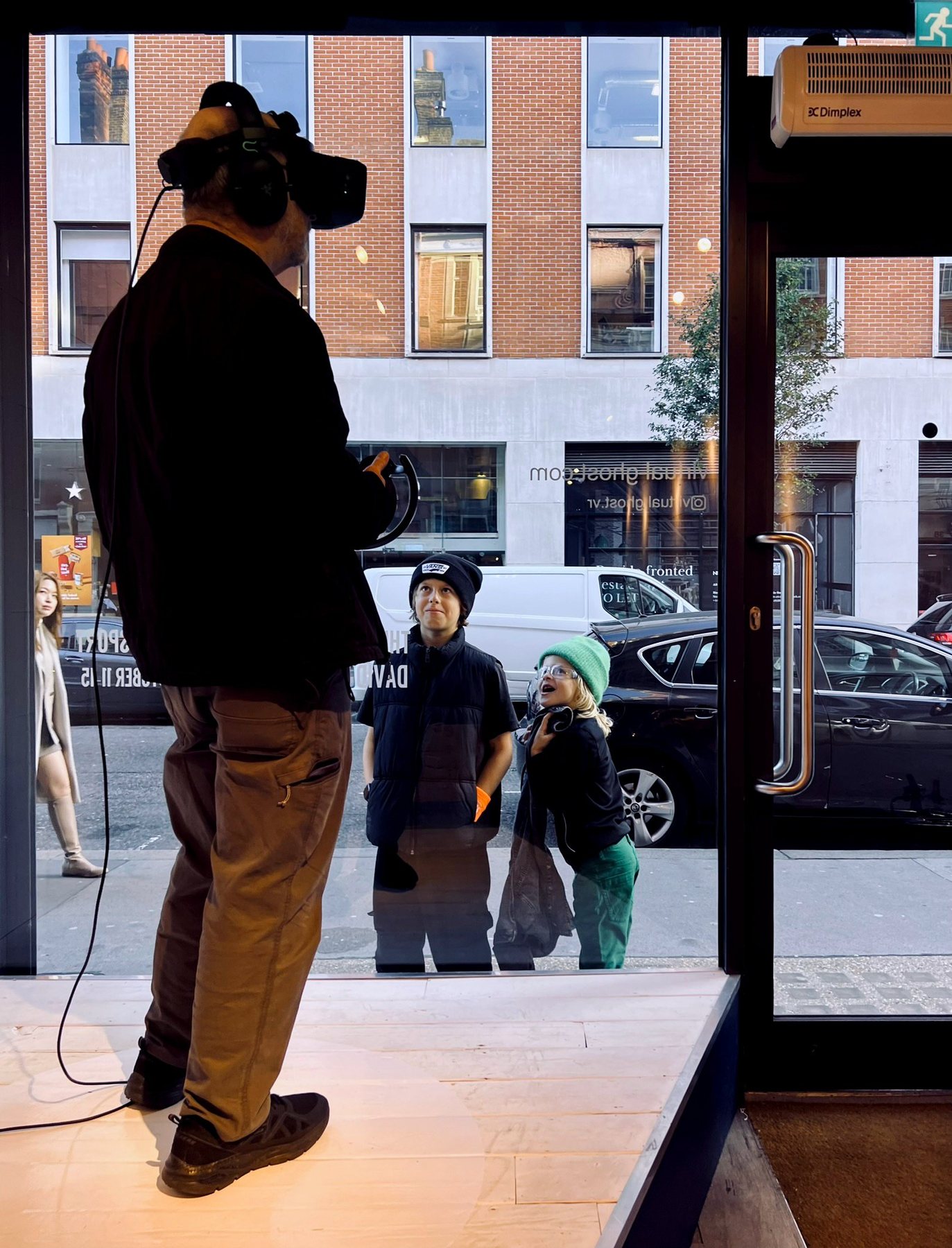 A man wearing a headset stands in the gallery window while two children look in from the street.
