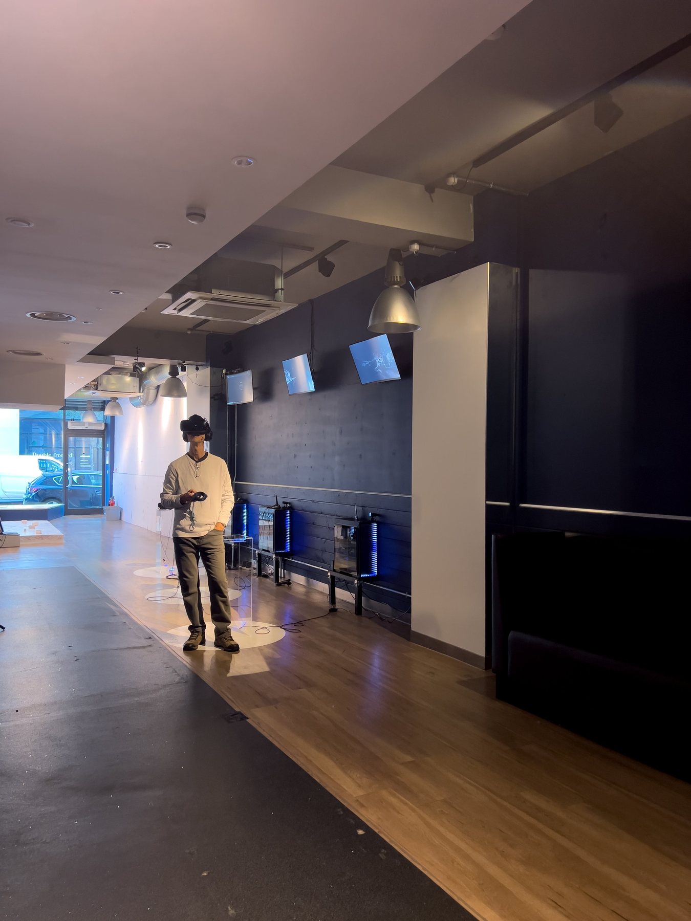 A man in a headset holding a controller stands in the dim gallery, with screens and clear plinths along the wall.