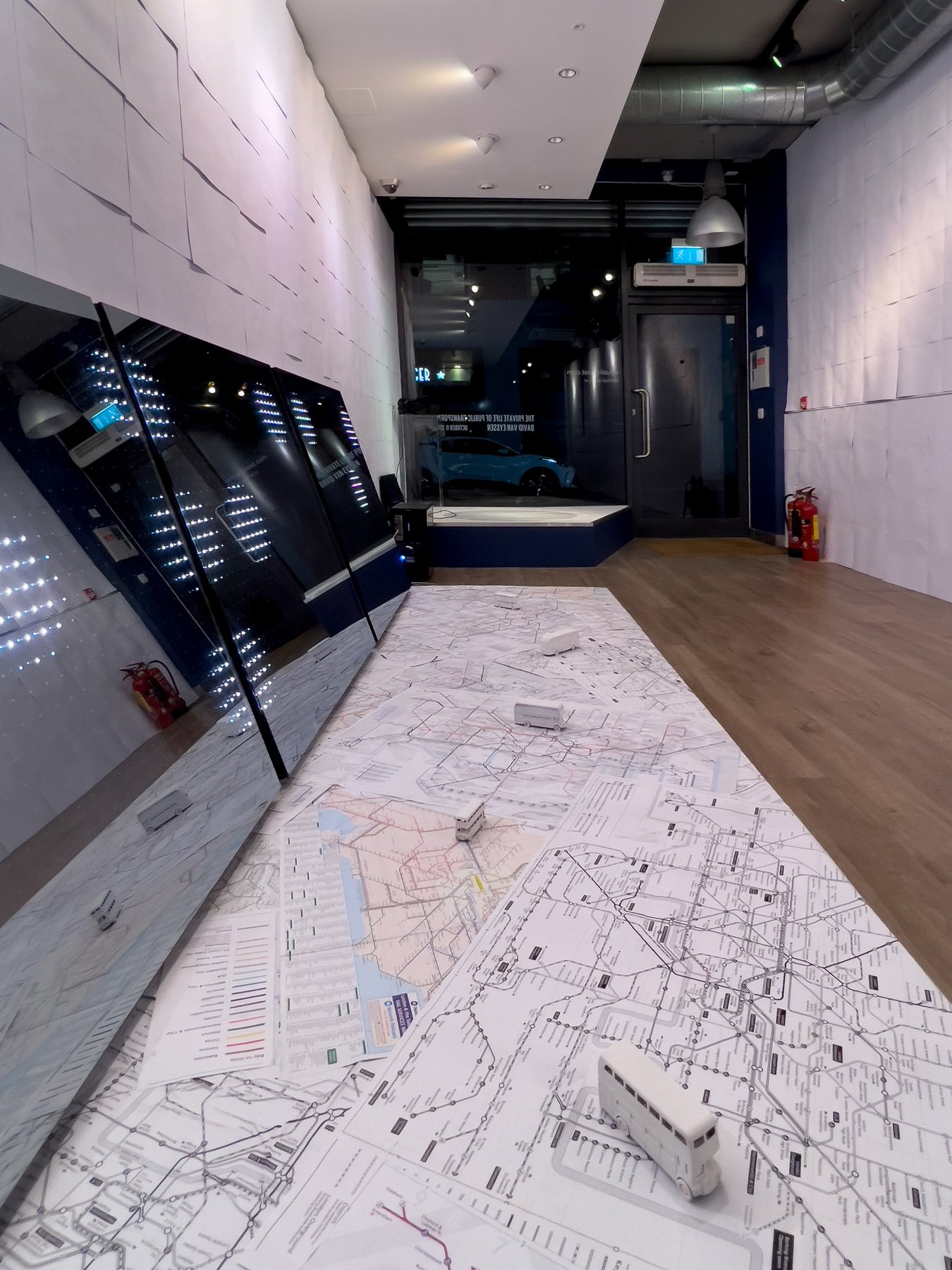 Floor map of London transport routes with white miniature buses, leading toward the gallery entrance beside a mirrored panel.