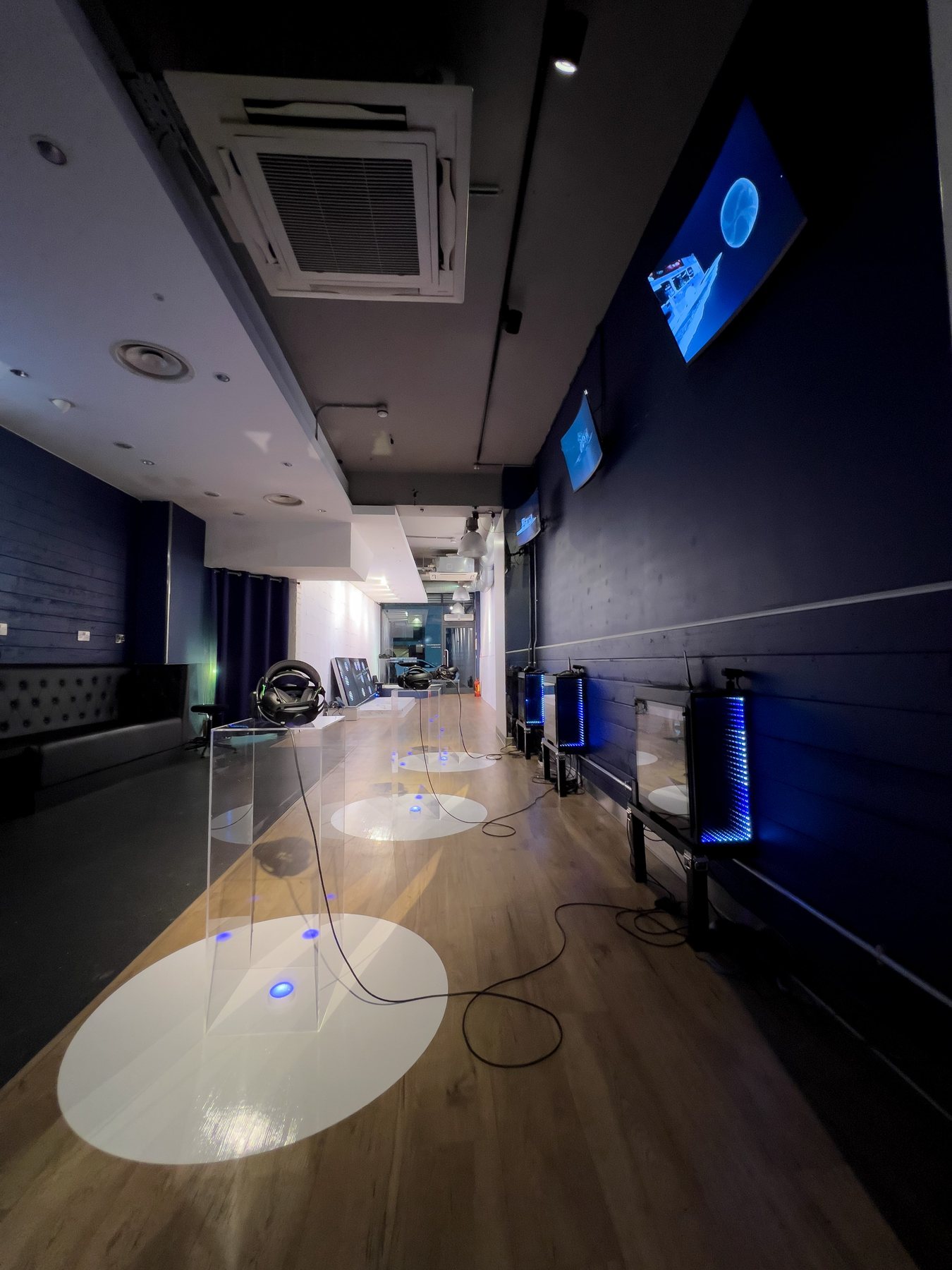Long dark-blue gallery with clear plinths on circular light pools, each holding a headset, and screens on the wall.