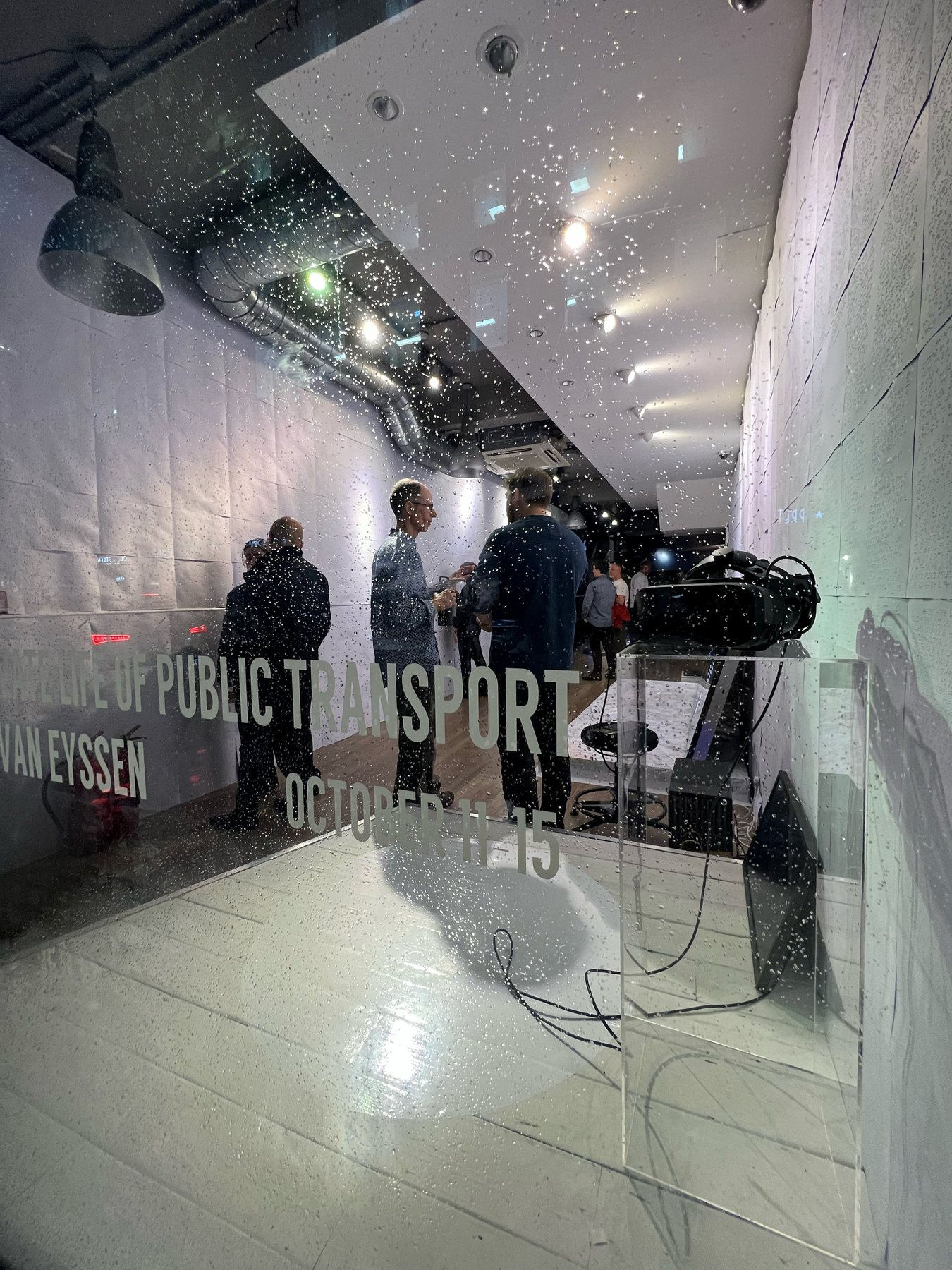 Gallery window etched with the exhibition title and dates, visitors reflected inside beside a headset on a clear plinth.