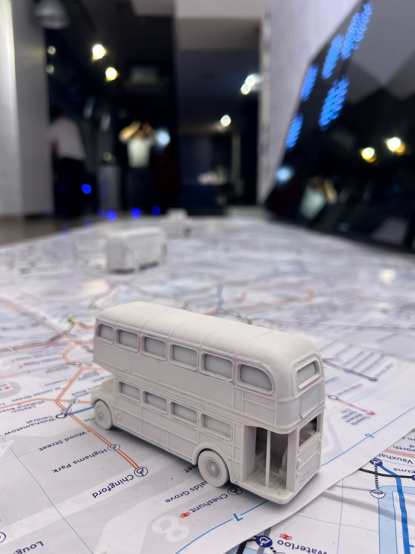 White miniature double-decker bus on a floor-sized London transport map, with visitors blurred in a dim gallery behind.