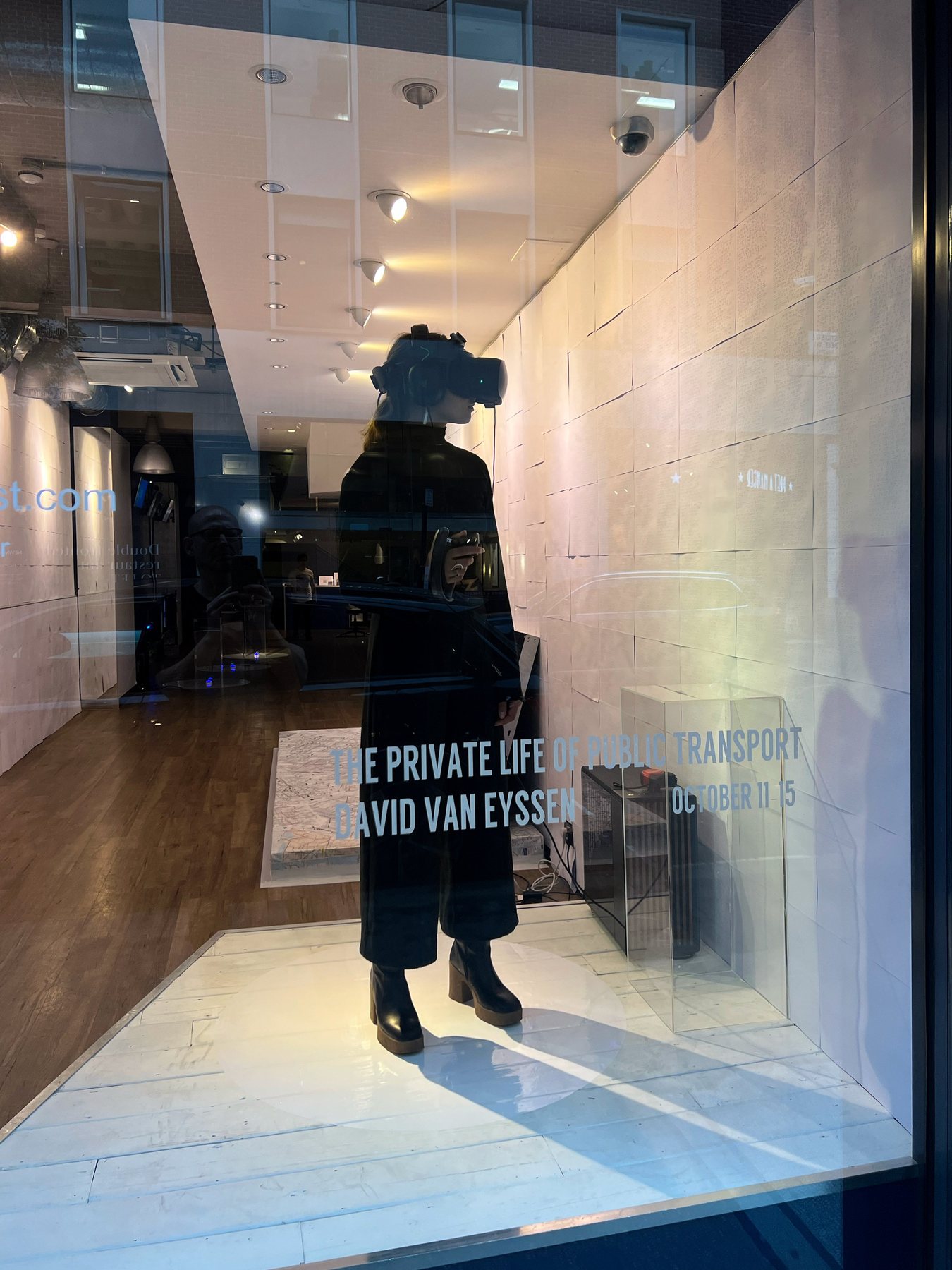 A dark-clothed visitor wearing a headset stands on a white platform inside the shopfront window, with the exhibition title on the glass.