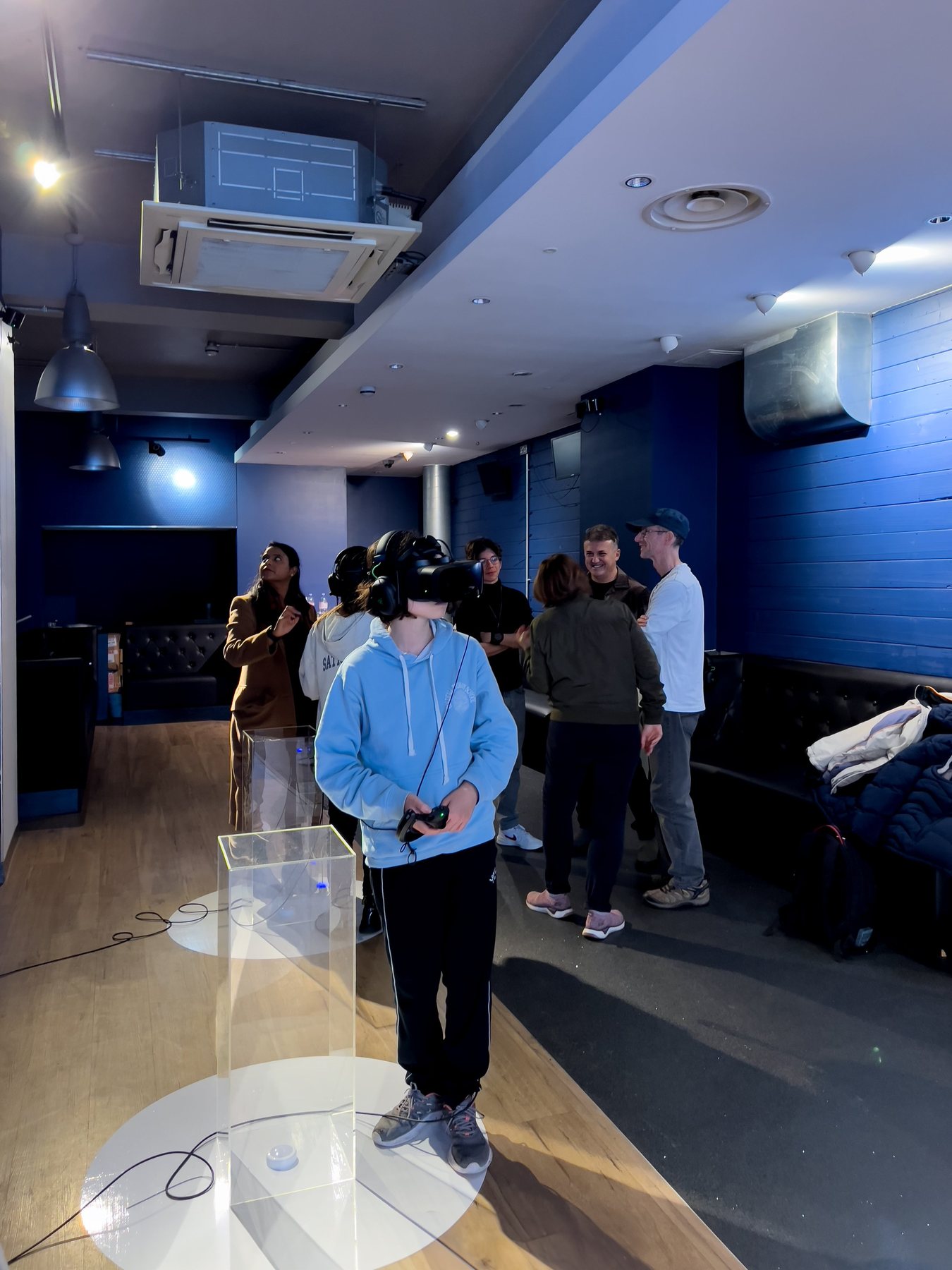 A visitor in a light-blue hoodie wearing a headset and holding a controller beside a clear plinth, with a small crowd behind.