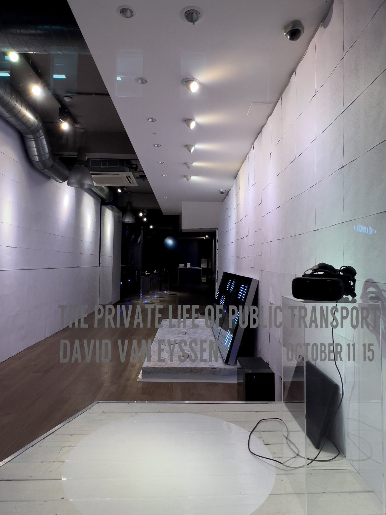 Long white gallery with braille wallpaper, a map-covered platform and a headset on a plinth, with the exhibition title overlaid.