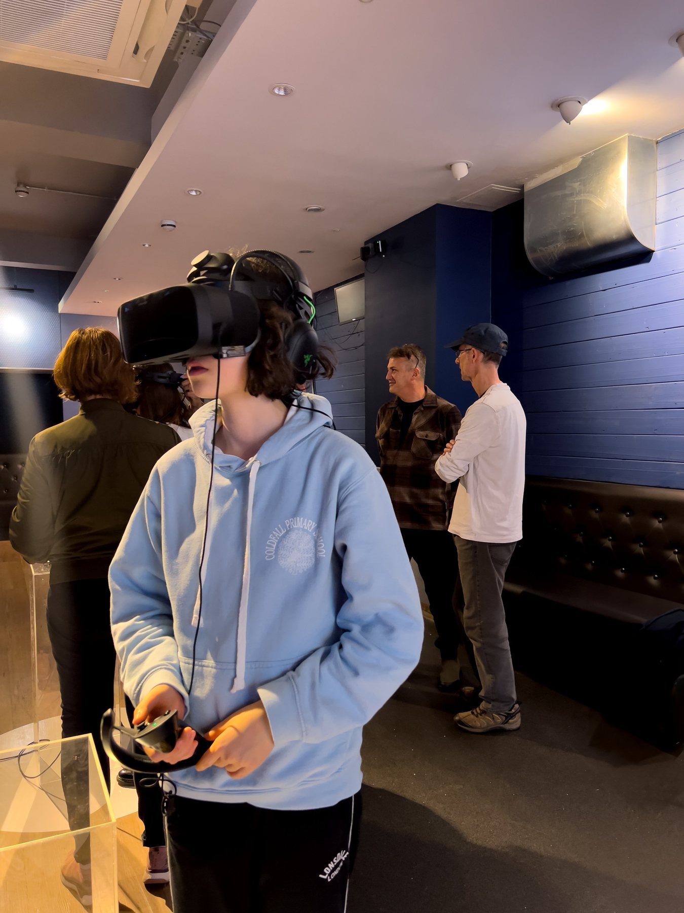 A visitor in a light-blue hoodie wearing a headset, holding a controller, in the blue-walled gallery with people behind.
