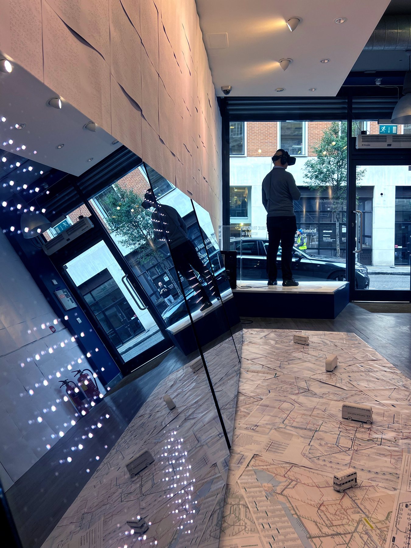 A visitor in a headset stands at the window end of the gallery, with a mirrored panel and miniature buses on a map floor.