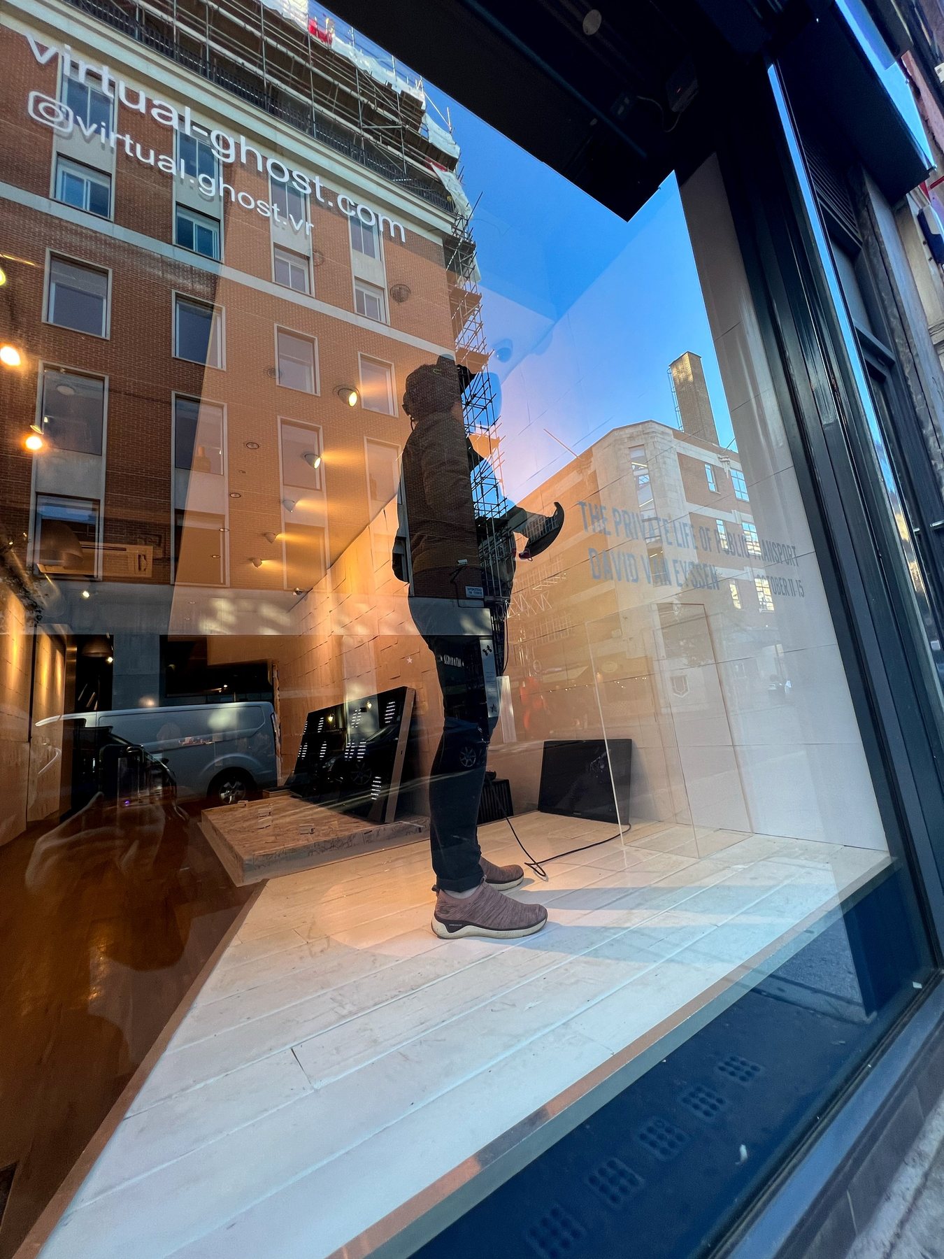 A person holding a controller stands inside the sunlit gallery window, the title and virtual-ghost.com reflected in the glass.