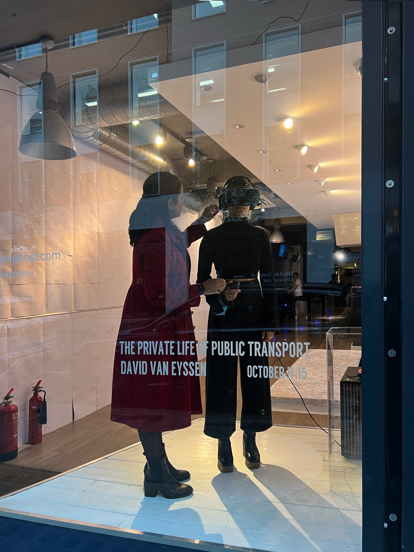 Two women at the gallery window, one helping the other adjust a headset, the title and dates on the glass.