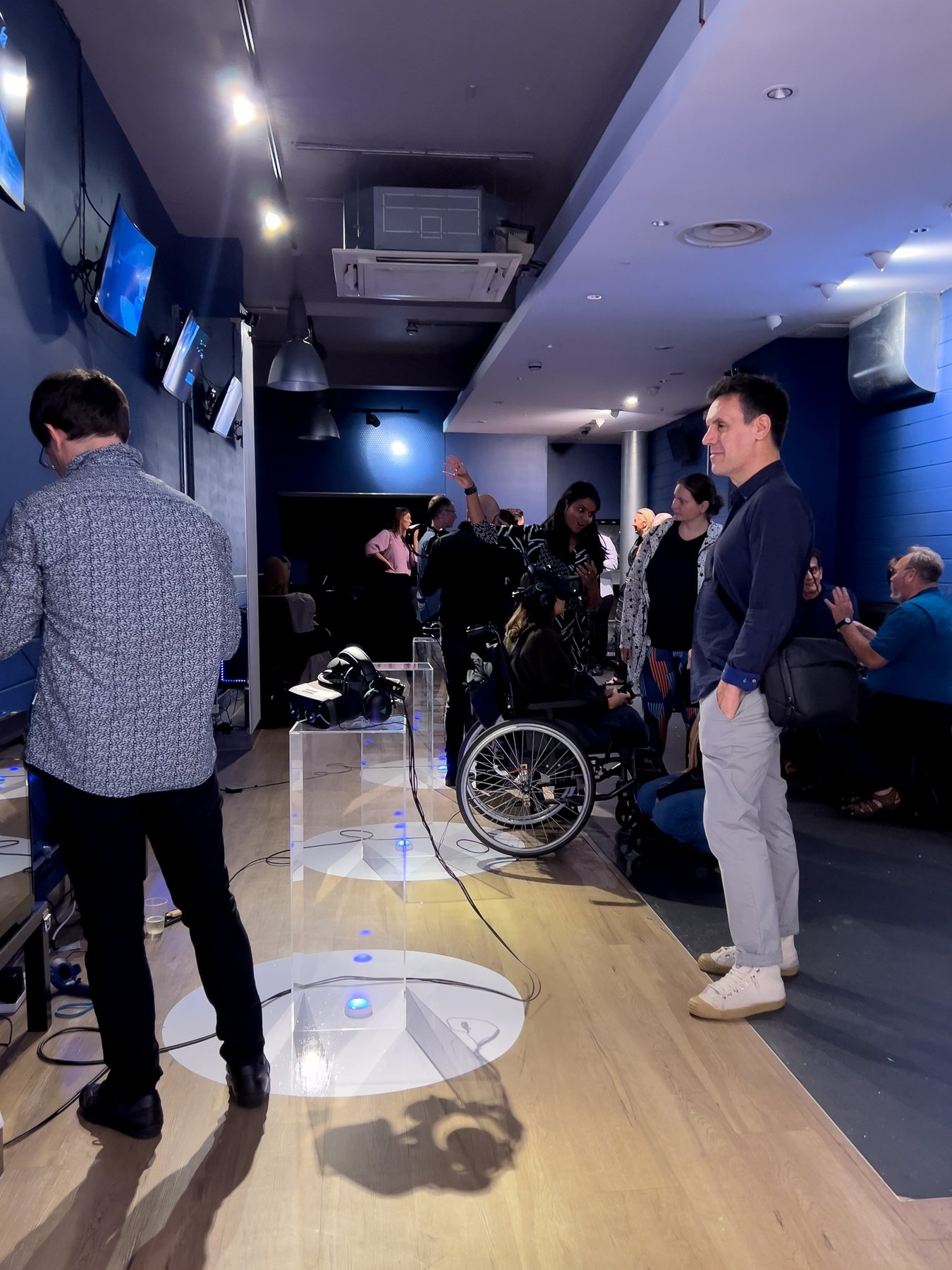 Visitors gathered in the blue-walled gallery, including a wheelchair user, around a clear plinth holding a headset.