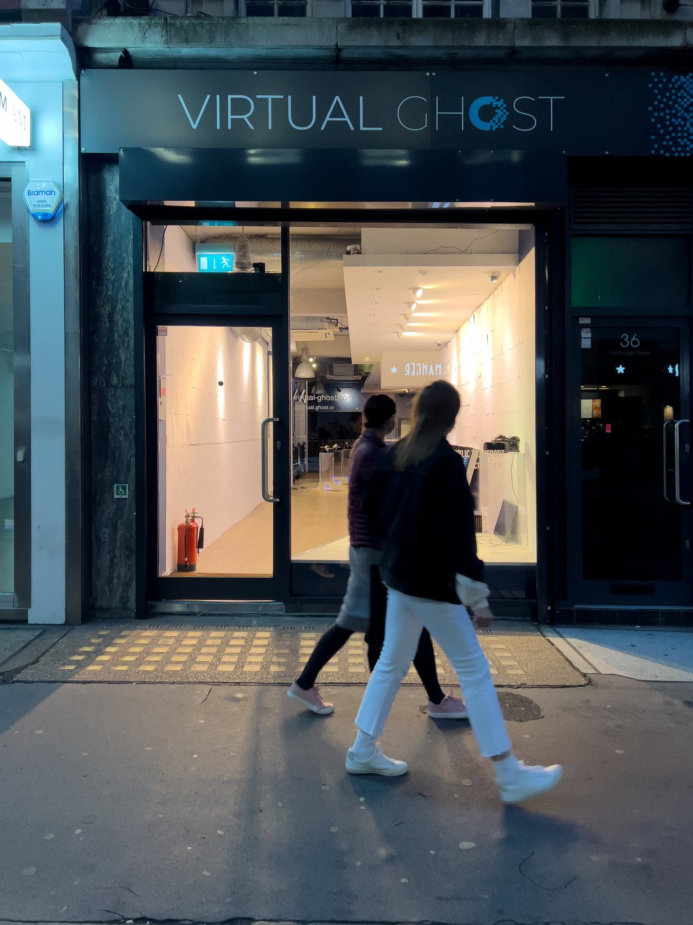 Shopfront of Virtual Ghost at dusk, with two people walking past the lit white gallery interior.