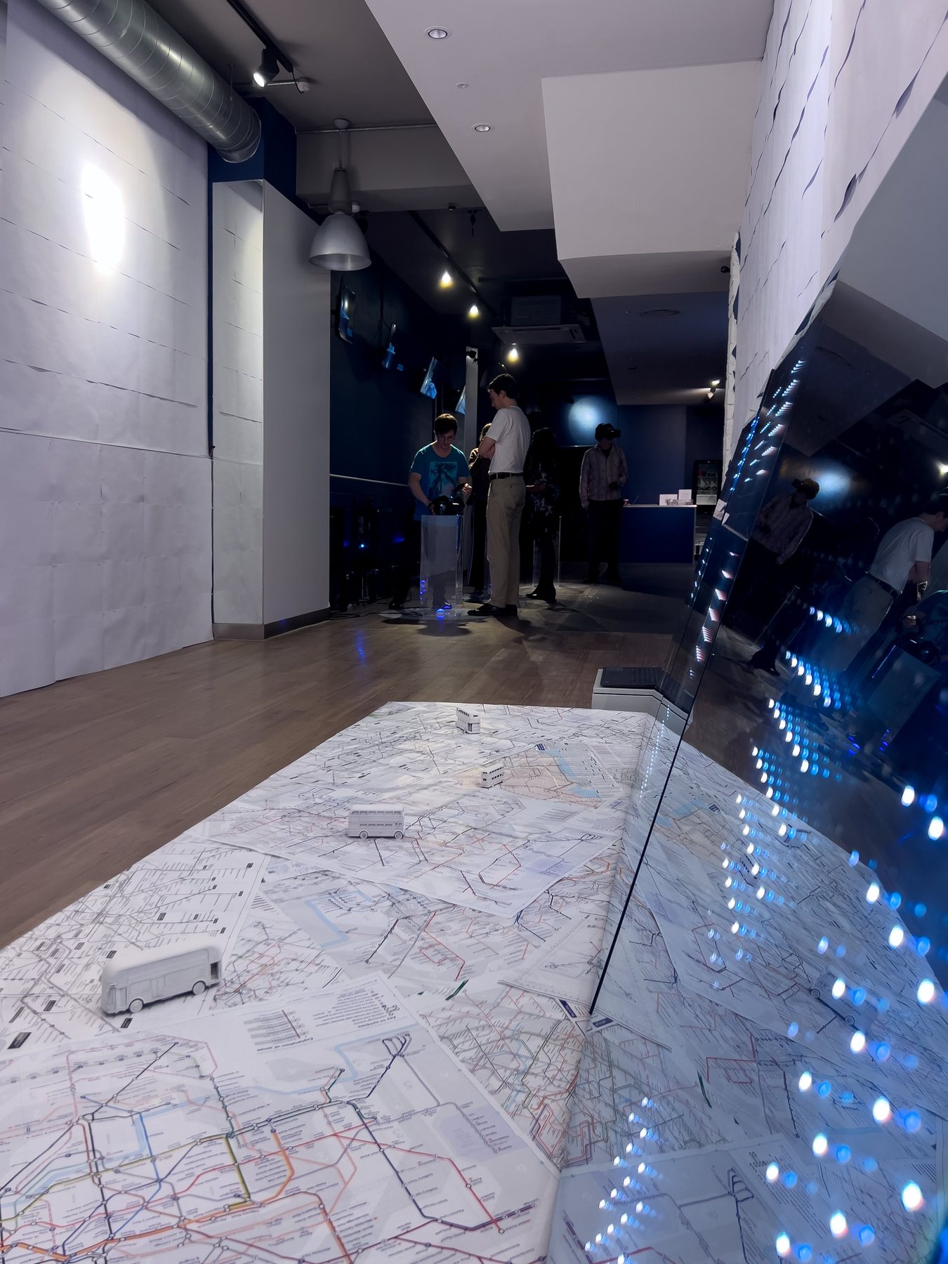 Visitors in the dim gallery beyond a floor map with white miniature buses and a mirror panel of lights.
