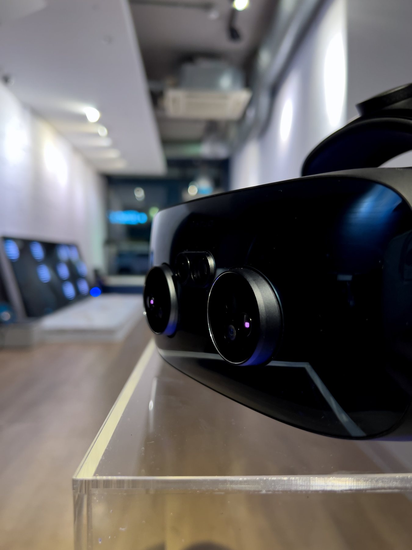 Close-up of a Varjo headset on a clear plinth, with the long white gallery blurred behind it.