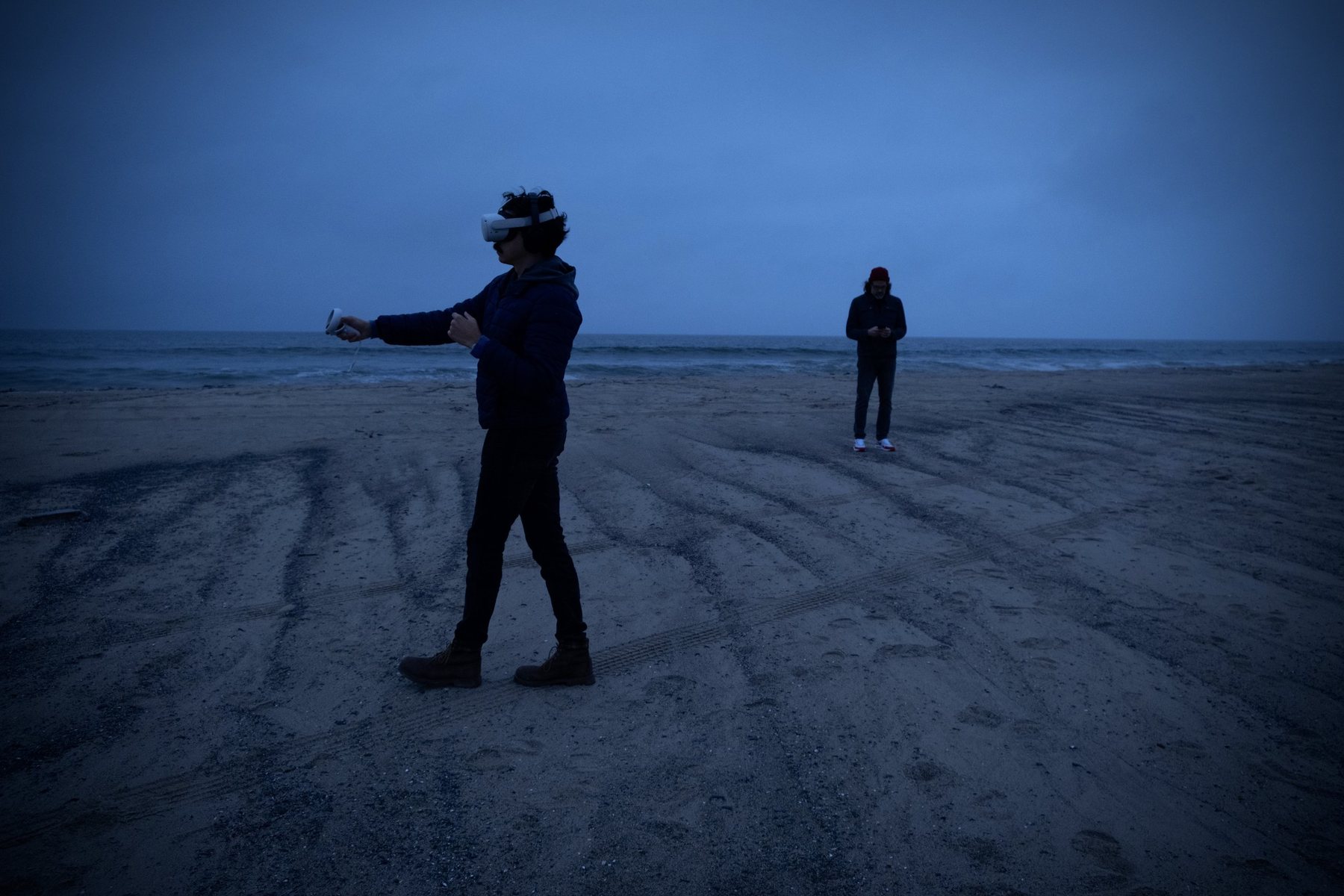 Two people in VR headsets on a wide dusk beach, one in the foreground reaching an arm out, the other standing farther away.