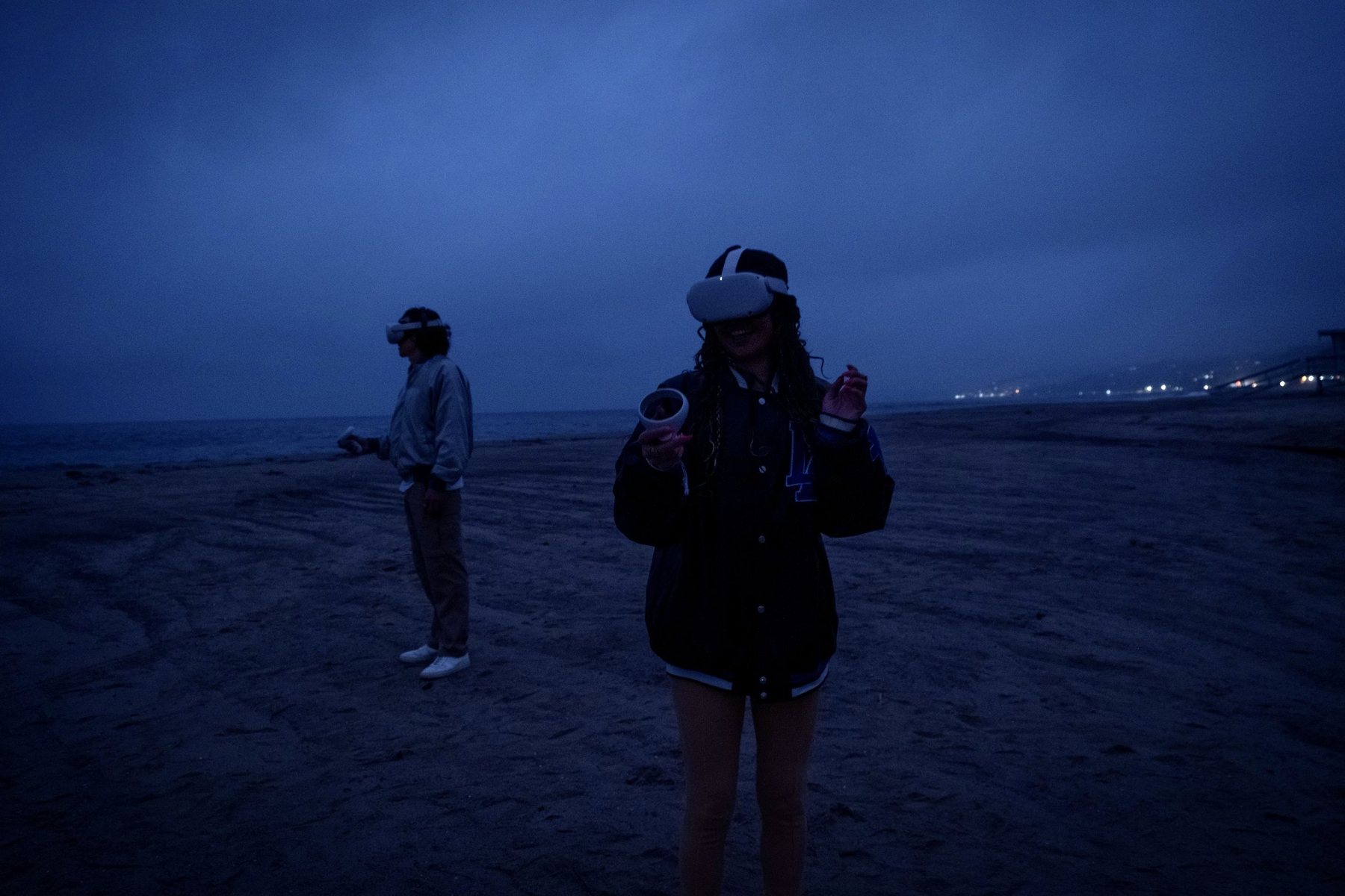 Two people in VR headsets hold controllers on a beach at dusk, the shoreline and lights behind them.