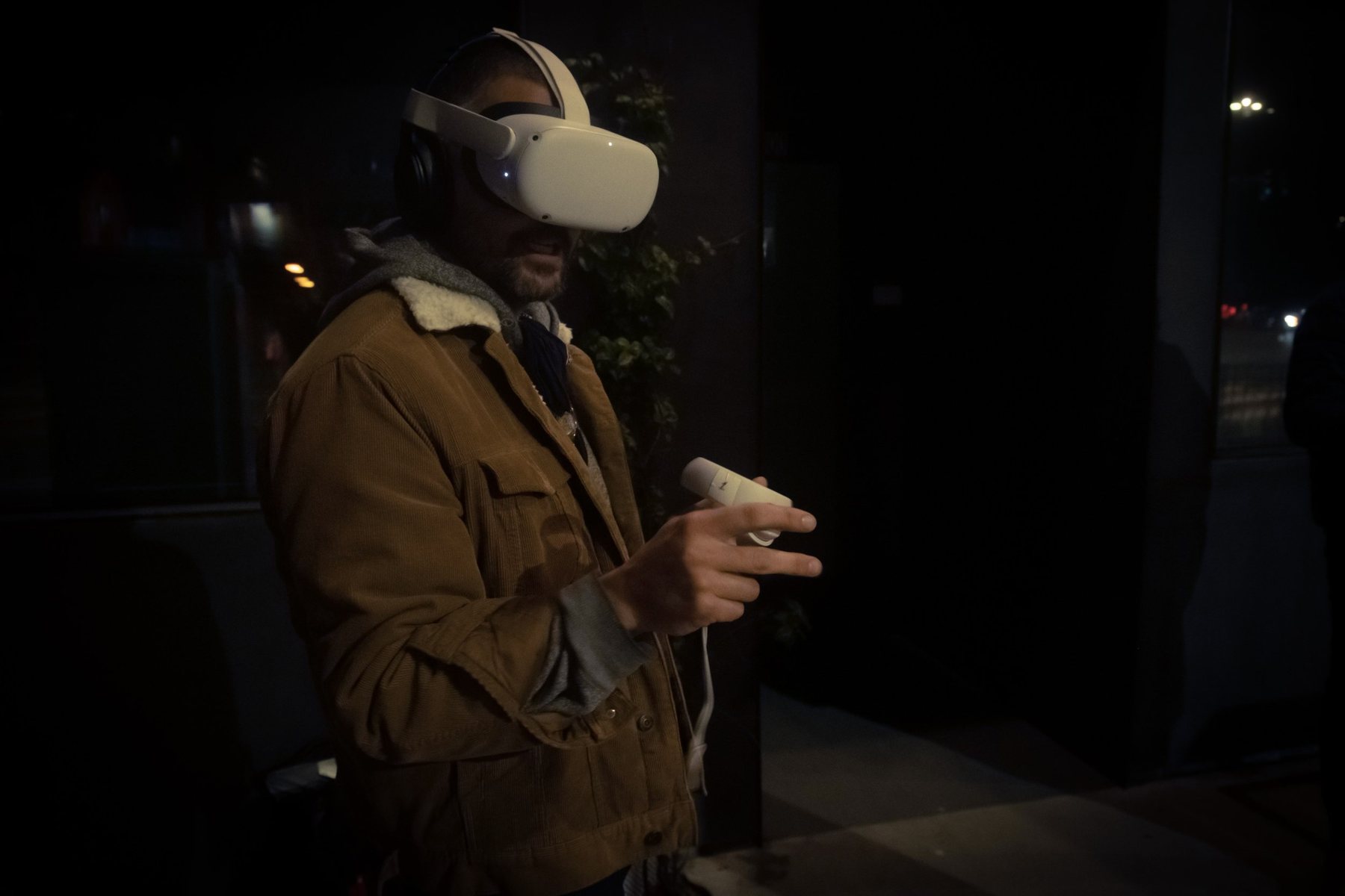 Man in a tan shearling-collar jacket wearing a white VR headset and holding a controller, standing in a dark night street setting.