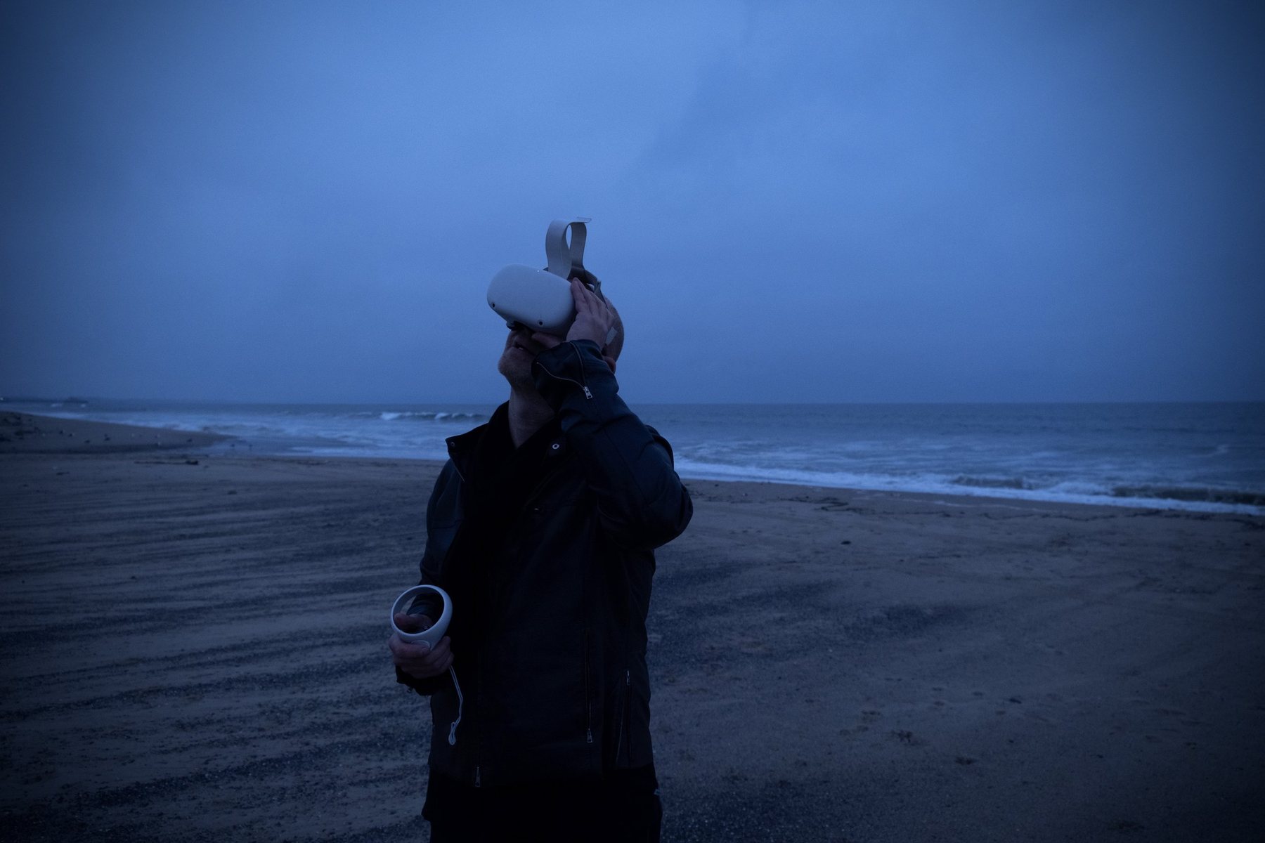 Person looking upward in a VR headset, controller in hand, on a dusky beach with grey surf under a deep blue sky.