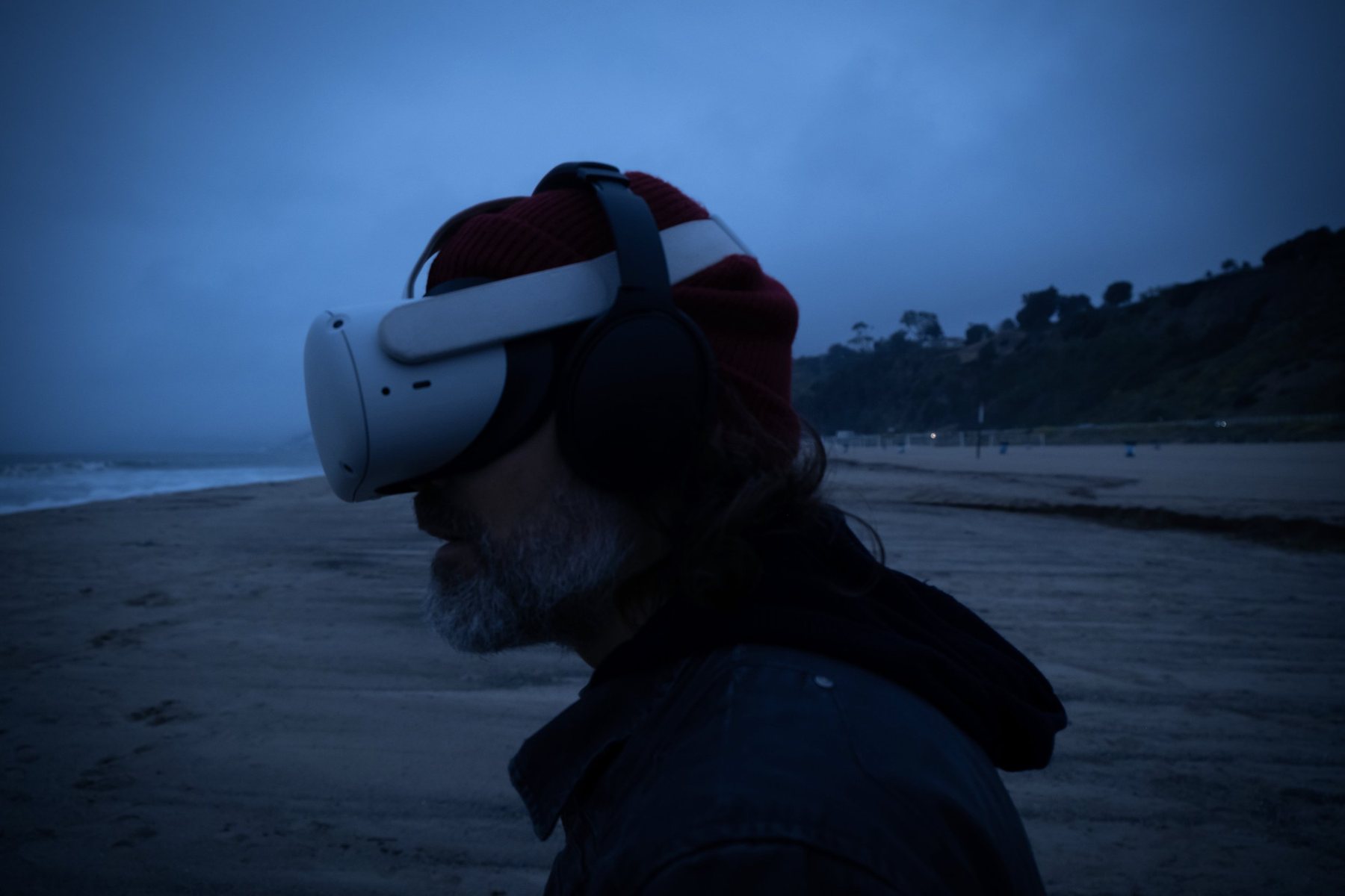 Profile of a bearded man in a red beanie, headphones and VR headset on a beach under a deep blue evening sky.