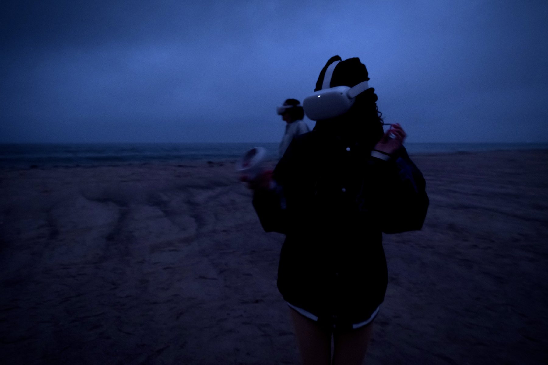 Woman in a dark jacket wearing a VR headset on a twilight beach, another headset wearer standing behind her.