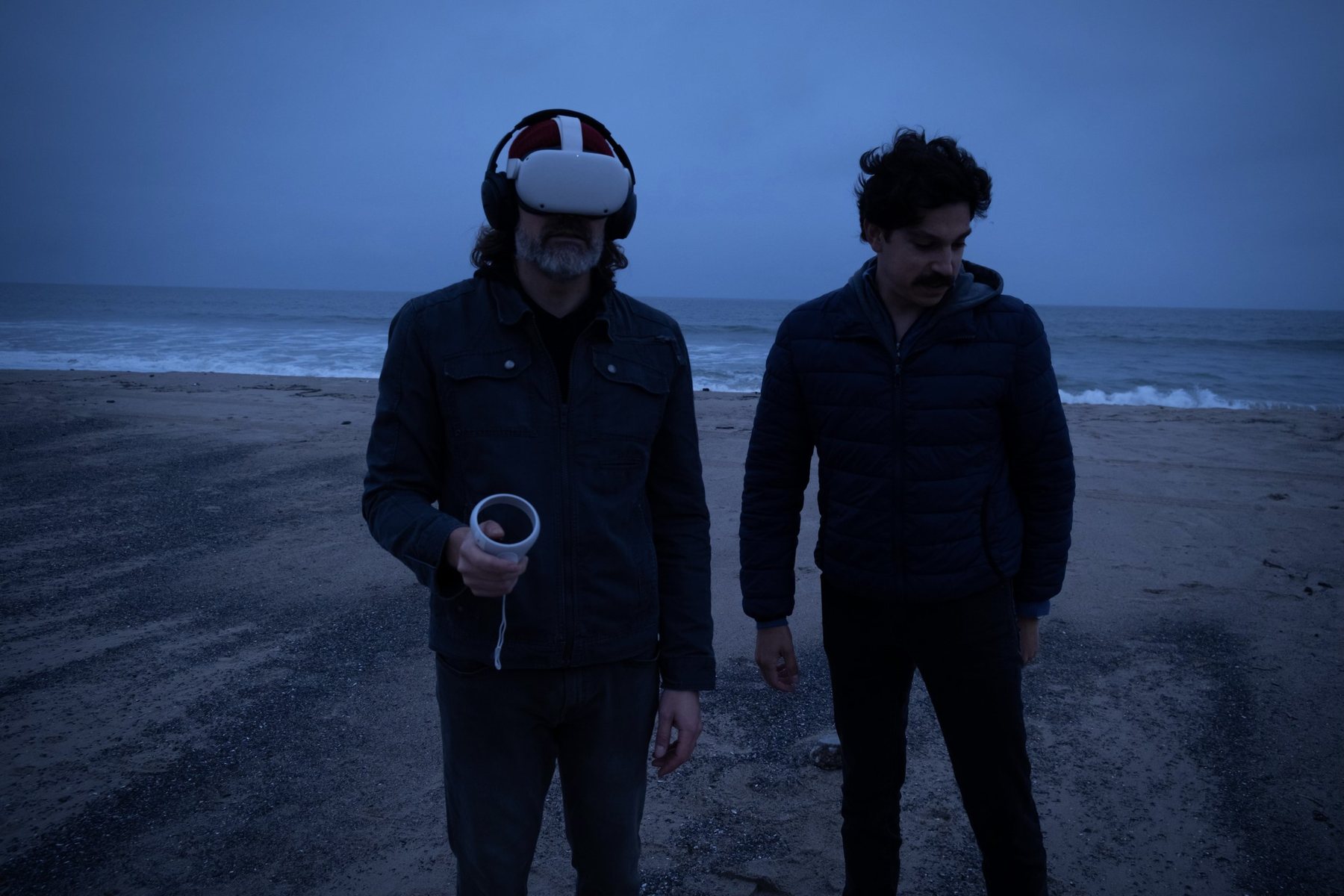 Two men on a beach at dusk, one wearing a VR headset and holding a controller, the other standing beside him.